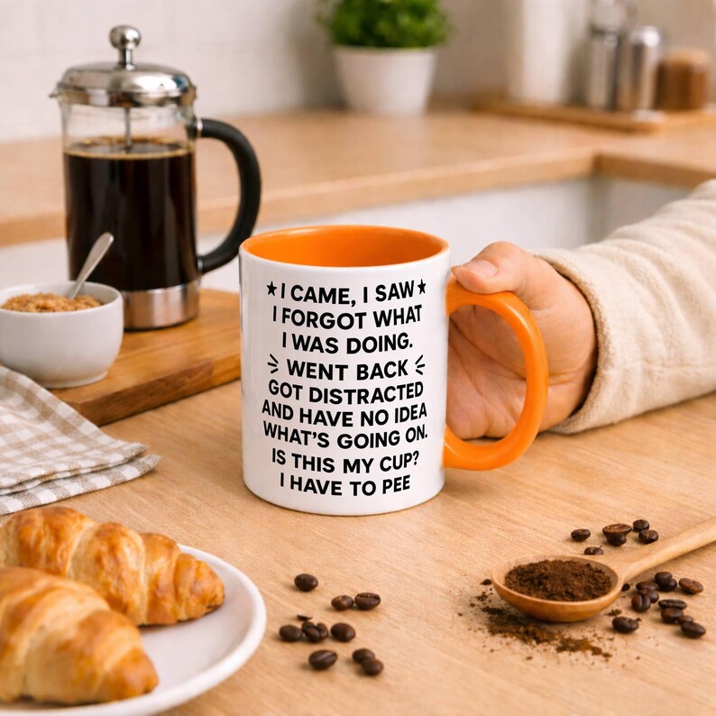 K&ouml;nnte beinhalten: Wei&szlig;er Kaffeebecher mit orangefarbenem Innenraum und Henkel, mit dem Text "I CAME, I SAW, I FORGOT WHAT I WAS DOING". Eine French Press, Croissants und Kaffeebohnen sind ebenfalls auf einer Holzoberfl&auml;che zu sehen.