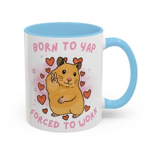May include: White ceramic mug with a light blue interior and handle. The mug features a cartoon hamster with a peace sign, surrounded by hearts, and the text "BORN TO YAP FORCED TO WORK" in pink.