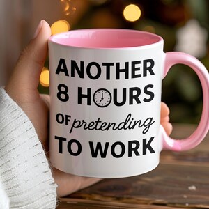 K&ouml;nnte beinhalten: Wei&szlig;er Keramikbecher mit rosa Innenraum und Henkel. Der Becher tr&auml;gt den schwarzen Text "ANOTHER 8 HOURS of pretending TO WORK" mit einer Uhrgrafik. Der Becher wird von einer Person gehalten.