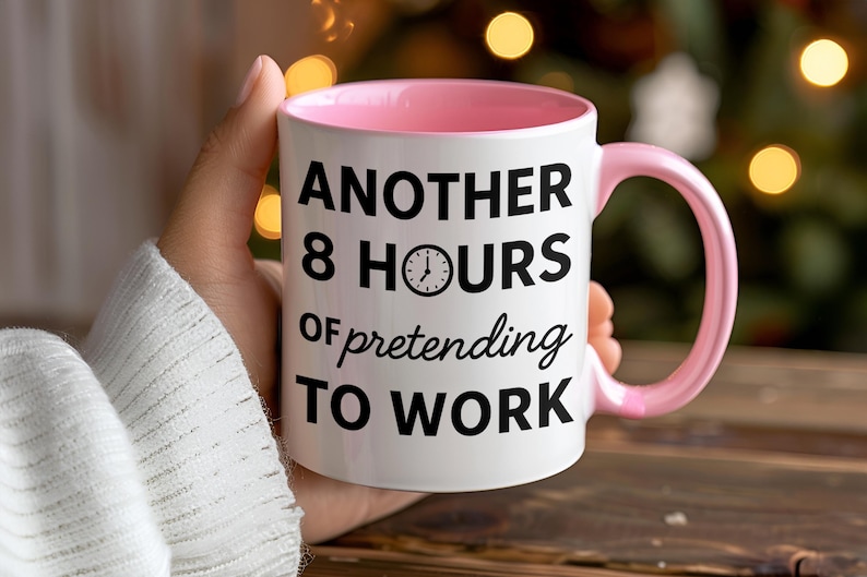 K&ouml;nnte beinhalten: Wei&szlig;er Keramikbecher mit rosa Innenraum und Henkel. Der Becher tr&auml;gt den schwarzen Text "ANOTHER 8 HOURS of pretending TO WORK" mit einer Uhrgrafik. Der Becher wird von einer Person gehalten.
