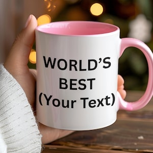 Puede incluir: Taza de cer&aacute;mica blanca con interior y asa rosas. La taza tiene el texto "WORLD'S BEST (Your Text)" en negro, perfecta para personalizar. La taza se sostiene en una mano, con un fondo borroso de luces.