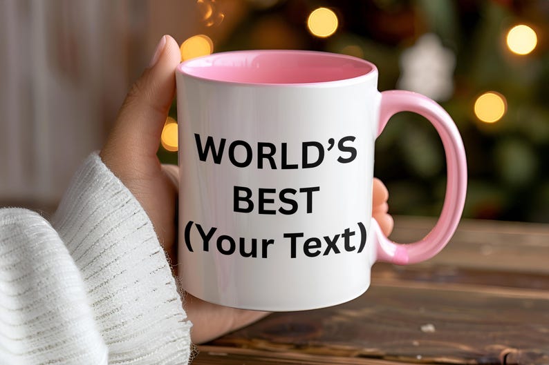 Puede incluir: Taza de cer&aacute;mica blanca con interior y asa rosas. La taza tiene el texto "WORLD'S BEST (Your Text)" en negro, perfecta para personalizar. La taza se sostiene en una mano, con un fondo borroso de luces.
