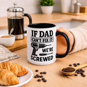 May include: White ceramic mug with a black interior and handle. The mug features the text "IF DAD CAN'T FIX IT WE'RE SCREWED" along with a graphic of a drill and screwdriver. The mug is held by a person.