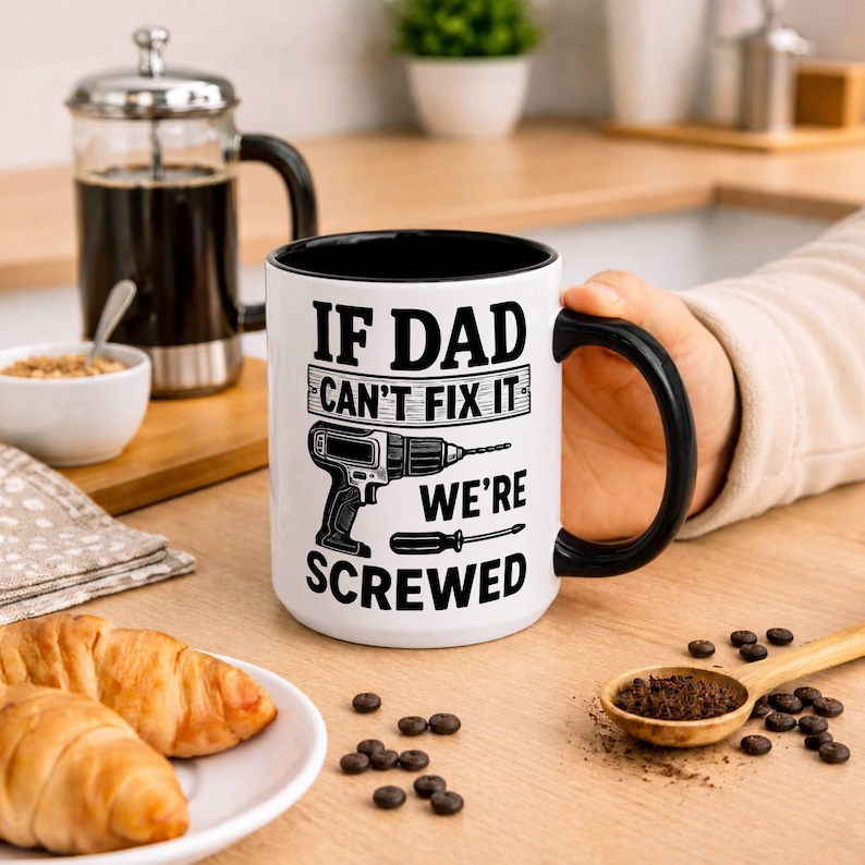 May include: White ceramic mug with a black interior and handle. The mug features the text "IF DAD CAN'T FIX IT WE'RE SCREWED" along with a graphic of a drill and screwdriver. The mug is held by a person.