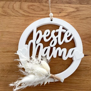 Könnte beinhalten: Ein weißer Holz-Kreis mit dem Text "beste mama" in einer dekorativen Schrift. Der Kreis hängt an einer Schnur und hat einen kleinen Strauß weißer Trockenblumen, der unten befestigt ist.