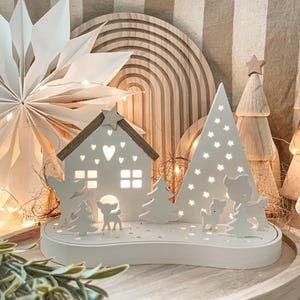 Könnte beinhalten: Weiße Weihnachtsdorf-Szene aus Holz mit einem Haus, zwei Bäumen und drei Rehen. Das Haus hat einen Stern oben und Herzen auf der Vorderseite. Die Bäume haben Sterne oben. Die Rehe stehen vor dem Haus und den Bäumen. Die Szene ist mit warmweißen Lichtern beleuchtet.