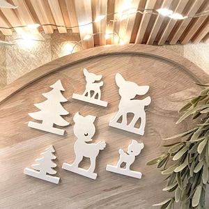 Könnte beinhalten: Ein Set aus sechs weißen Holz-Ausschnitten von Hirschen und Kiefern. Die Ausschnitte sind auf einer Holzoberfläche angeordnet.