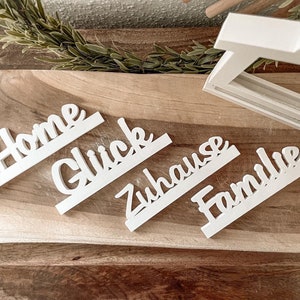 Könnte beinhalten: Vier weiße Holzschilder mit den Worten "Home", "Glück", "Zuhause" und "Familie" in dekorativer Schrift.