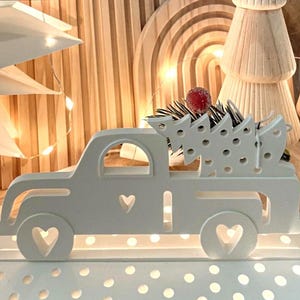 Könnte beinhalten: Weißer Holzlaster mit einem weißen Holz-Weihnachtsbaum in der Ladefläche. Der Laster hat ausgeschnittene Herzen an den Seiten und Rädern. Der Laster steht auf einem weißen Sockel mit einem Tupfenmuster.