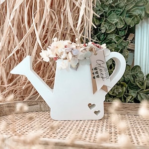 Könnte beinhalten: Weiße Keramik-Gießkanne mit Henkel und Ausguss. Die Kanne ist mit Trockenblumen gefüllt und hat ein kleines Schild am Henkel, auf dem "Ein Stück Glück" steht, was "Ein Stück Glück" bedeutet.