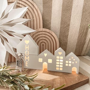 Könnte beinhalten: Weißes, hölzernes Haus in Form einer Leuchte mit einem 'Home'-Ausschnitt und einem kleinen Etikett mit der Aufschrift 'The most important thing is Glück'. Die Leuchte ist beleuchtet und steht auf einer Holzoberfläche.