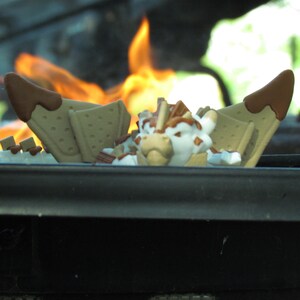 May include: A whimsical s'mores-themed decorative item. The design features graham crackers with chocolate and marshmallow elements. The background includes a blurred view of flames, suggesting a campfire setting.