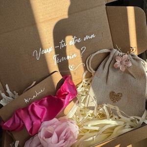 Può includere: Una scatola regalo con un messaggio scritto a mano in grafia bianca, un elastico rosa, un fiore rosa e una piccola borsa beige con un cuore dorato. La scatola contiene carta decorativa e un anello d'oro.