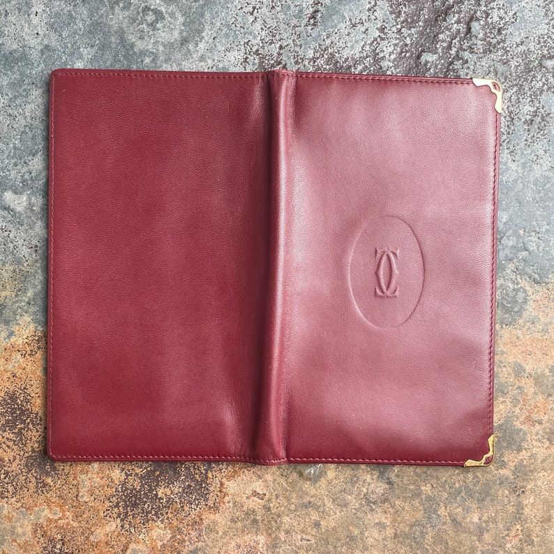 May include: A burgundy leather wallet with gold accents and the Cartier logo embossed on the front.