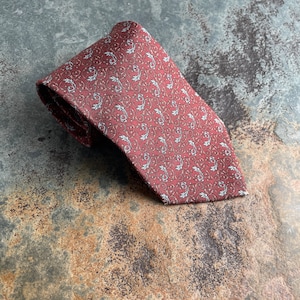 May include: A folded burgundy tie with a repeating pattern of white and light brown stylized leaves and vines. The tie is made of a smooth fabric and is displayed on a textured, multi-toned surface.