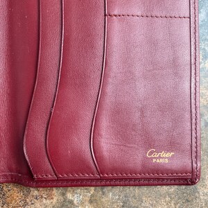 May include: A burgundy leather wallet with a stitched edge and the words 'Cartier PARIS' embossed in gold lettering.
