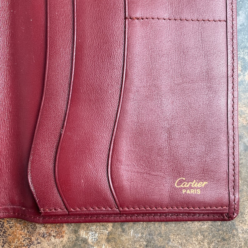 May include: A burgundy leather wallet with a stitched edge and the words 'Cartier PARIS' embossed in gold lettering.