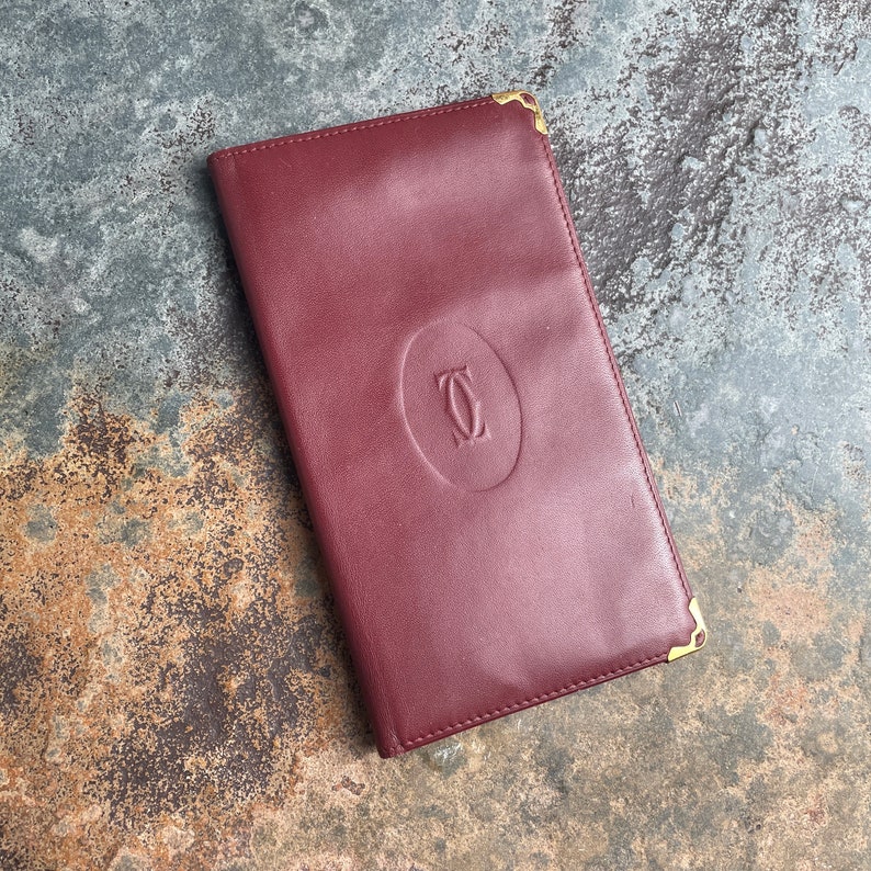 May include: A burgundy leather wallet with gold accents and the Cartier logo embossed on the front.
