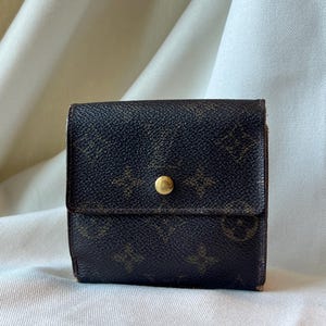 May include: A small, square, dark brown wallet with a gold-colored snap closure. The wallet features a repeating pattern of brown logos and symbols. The wallet is closed and sits on a white surface.