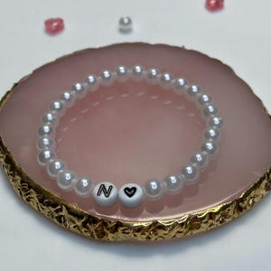 Könnte beinhalten: Ein weißes Perlenarmband mit dem Buchstaben "N" und einem Herzanhänger. Das Armband besteht aus kleinen, runden, weißen Perlen. Es liegt auf einer rosa-goldenen Oberfläche.