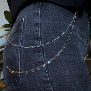Könnte beinhalten: Eine silberfarbene Taillenkette mit zwei Lagen, bestehend aus einer einfachen Gliederkette oben und einer zweiten Kette, die mit kleinen, gleichmäßig verteilten metallischen Sternanhängern verziert ist, getragen über einer dunkelgrauen Jeans.