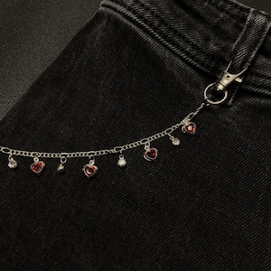 Könnte beinhalten: Eine silberfarbene Metallketten-Accessoire, das mit einem Karabinerverschluss an einem schwarzen Jeansstoff befestigt ist. Die Kette ist mit abwechselnden Anhängern versehen, darunter kleine rote herzförmige Steine und klare runde Kristalle.