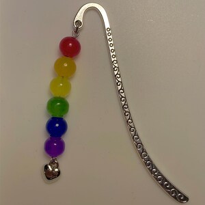 May include: A silver bookmark with a curved design and small circles. It has a strand of rainbow-coloured beads in red, yellow, green, blue, and purple, and a small silver heart charm.
