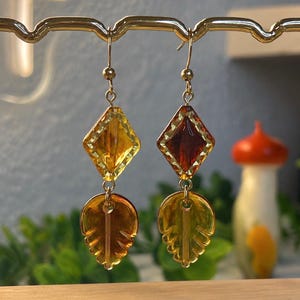May include: A pair of gold-toned dangle earrings featuring a faceted diamond-shaped bead in yellow and deep amber, suspended above a translucent amber-colored leaf-shaped charm. The earrings hang from gold-toned metal fishhook wires.