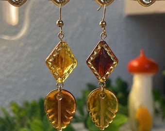 May include: A pair of gold-toned dangle earrings featuring a faceted diamond-shaped bead in yellow and deep amber, suspended above a translucent amber-coloured leaf-shaped charm. The earrings hang from gold-toned metal fishhook wires.