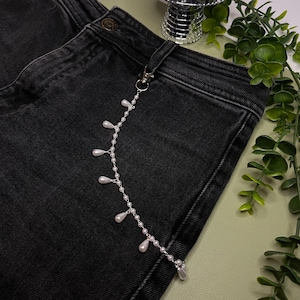 May include: A silver-toned metal chain attached to a belt loop of black denim jeans. The chain features a series of small white spherical beads interspersed with dangling white teardrop-shaped faux pearls along its entire length.