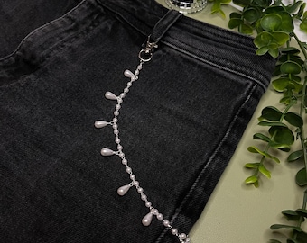 May include: A silver-toned metal chain attached to a belt loop of black denim jeans. The chain features a series of small white spherical beads interspersed with dangling white teardrop-shaped faux pearls along its entire length.