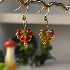 Könnte beinhalten: Ein Paar Ohrhänger mit bernsteinfarbenen, blattförmigen Glasanhängern. Jeder Ohrring hängt an einem goldfarbenen Metallhaken und ist mit einer kleinen goldfarbenen Perle über dem Blattanhänger sowie einer kleineren Perle am unteren Ende versehen.