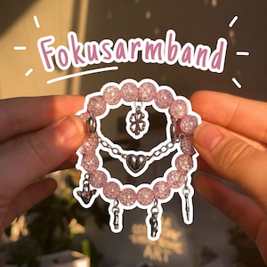 Könnte beinhalten: Ein Armband aus hellrosa Crackle-Glasperlen mit silberfarbenen Anhängern, darunter ein vierblättriges Kleeblatt, ein Herz und verschiedene kleine Anhänger. Das Wort Fokusarmband steht in rosa Schrift über dem Armband.