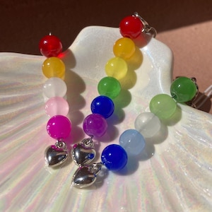 Könnte beinhalten: Regenbogenfarbene Perlenohrringe mit silbernen Herzanhängern. Die Ohrringe zeigen einen Farbverlauf aus roten, gelben, weißen, rosa, magenta, lila, blauen, grünen und hellblauen Perlen. Die Ohrringe werden auf einer weißen, irisierenden Oberfläche präsentiert.