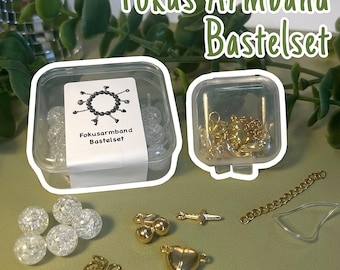 Könnte beinhalten: Ein DIY-Armband-Bastelset mit der Aufschrift Fokus Armband Bastelset, bestehend aus zwei transparenten Kunststoffbehältern, klaren Crackle-Glasperlen, goldfarbenen Metallanhängern in Herz- und Kirschform, Biegeringen, einer Verlängerungskette und elastischem Band.