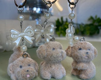 Puede incluir: Tres llaveros con figuras de perro de peluche marrón claro, cada uno con un lazo blanco o detalle de perla. Los llaveros tienen anillos de metal plateado y cuentas de perlas. Los perros tienen ojos negros y lenguas rosadas.