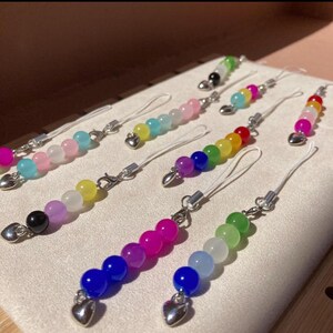 May include: A selection of beaded phone charms in a variety of colours. Each charm has a string of round beads in shades like pink, blue, green, and yellow. The charms have a silver heart-shaped charm and a silver clasp.