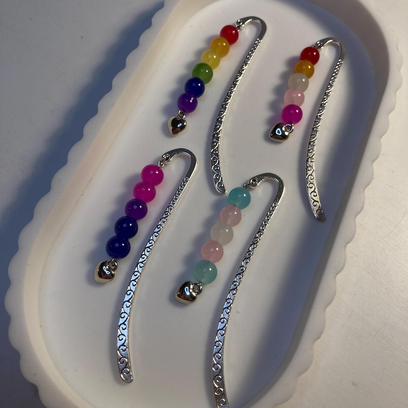 May include: A selection of beaded bookmarks with silver metal detailing. Each bookmark features a different colour scheme of beads, including rainbow, pink, and blue. The bookmarks have a heart-shaped charm at the bottom.