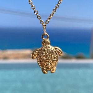 May include: A gold-colored necklace featuring a turtle pendant. The pendant is intricately detailed with a textured surface and a small loop for the chain. The chain is also gold and hangs against a blurred background of blue water and sky.