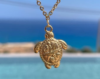May include: A gold-coloured necklace with a turtle pendant. The pendant has detailed, textured features and a small loop for the chain. The chain is gold and hangs against a blurred background of blue water and sky.