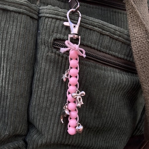 Könnte beinhalten: Ein rosa Perlen-Schlüsselanhänger mit silbernen Anhängern, darunter ein Stern, eine Schleife, ein Delphin und ein Herz. Der Schlüsselanhänger ist an einem silbernen Verschluss und einer weißen Schleife befestigt. Der Hintergrund ist ein dunkelgrüner Cordstoff.