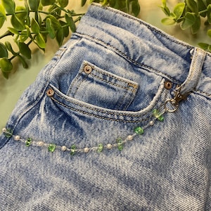Könnte beinhalten: Ein hellblauer Jeansstoff mit einer dekorativen Kette, die mit einem Metallkarabiner an einer Gürtelschlaufe befestigt ist. Die Kette besteht aus kleinen weißen Perlen, die mit unregelmäßigen, durchscheinenden grünen Steinen durchsetzt sind.