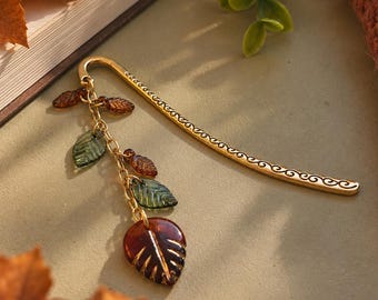 Puede incluir: Un marcapáginas de metal dorado en forma de gancho con un patrón de volutas grabado a lo largo del cuerpo. Una cadena dorada está sujeta a la parte superior, decorada con cinco dijes de vidrio translúcido en forma de hojas en tonos ámbar, marrón y verde oscuro.