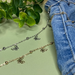 Könnte beinhalten: Zwei zarte Ketten, eine silberfarbene und eine goldfarbene, jeweils mit kleinen Metallperlen und dünnen, durchbrochenen Schmetterlingsanhängern. Die Ketten liegen auf einem blauen Jeansstoff und zeigen die filigrane Metallverarbeitung.