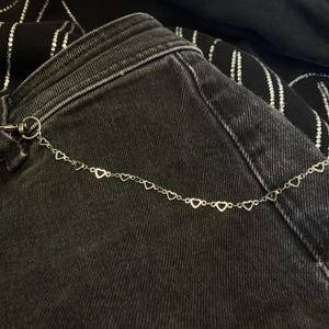 Könnte beinhalten: Eine silberfarbene Metallkette mit einer Reihe kleiner, offener Herzglieder, die an einem runden Verschluss befestigt ist. Die Kette liegt auf einer dunkelgrauen Jeans, die mit einem schwarzen Stoffüberzug mit vertikalen Strassstreifen versehen ist.