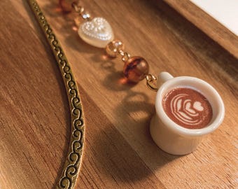 Könnte beinhalten: Goldfarbener Metall-Lesezeichen mit dekorativem Wirbeldesign. Der Lesezeichen hat einen Anhänger mit einer weißen herzförmigen Perle, bernsteinfarbenen Perlen und einer Miniatur-Kaffeetasse mit Latte Art. Auf einer Holzoberfläche.
