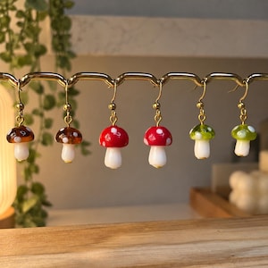May include: Six mushroom-shaped earrings hanging from a gold-toned metal display rack. The earrings feature white stems and caps in various colors including brown, red, and green, all decorated with small white dots and gold-toned hooks.