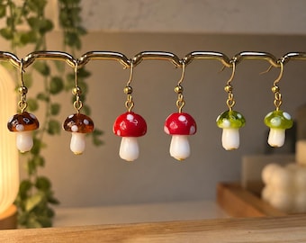 May include: Six mushroom-shaped earrings hanging from a gold-toned metal display rack. The earrings feature white stems and caps in various colours including brown, red, and green, all decorated with small white dots and gold-toned hooks.