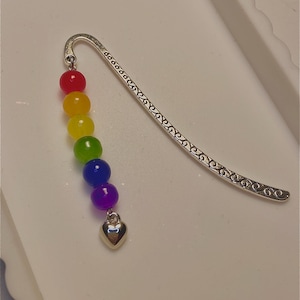 Könnte beinhalten: Ein silberner Lesezeichen mit dekorativem Design und einer Regenbogen-Perlenkette. Die Kette hat rote, gelbe, grüne, blaue und violette Perlen und endet mit einem kleinen silbernen Herzanhänger.