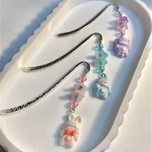 Könnte beinhalten: Drei silberne Metall-Lesezeichen mit dekorativen Anhängern. Die Anhänger umfassen ein weißes Kaninchen mit rosa Schleife, ein blaues Wesen und einen lila Bären. Die Anhänger sind mit rosa, blauen und lila Perlen an den Lesezeichen befestigt.